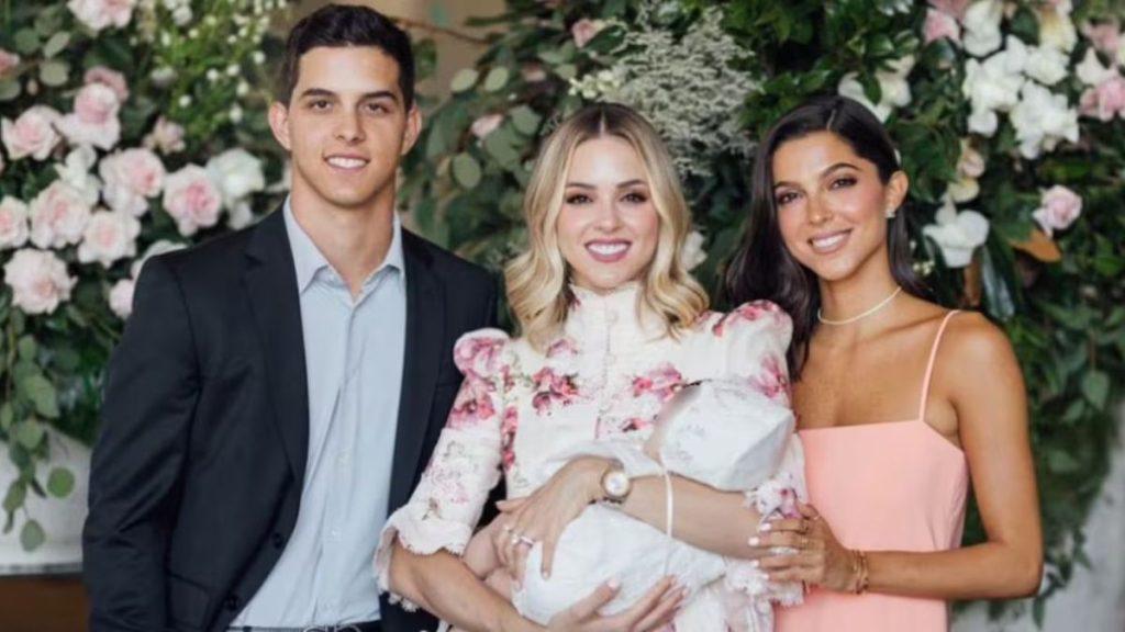 Three People Posing for a Family Photo in Front of a Flower Backdrop: a Man in a Suit, a Smiling Woman Holding a Baby, and a Woman in a Pink Dress. - Especies Políticas