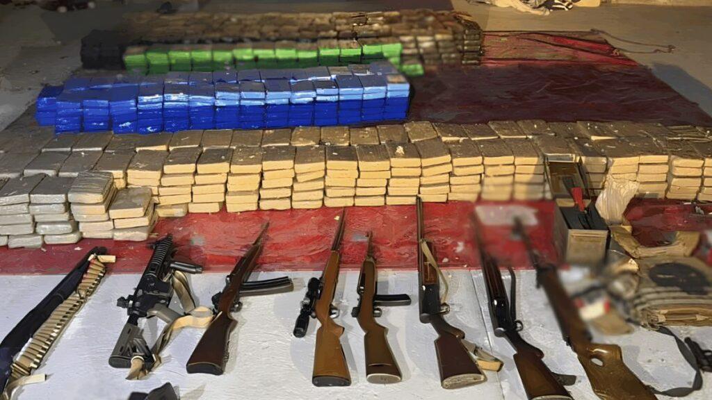 Bloque 2- Junio 3 - Especies Políticas Collection of Firearms and Ammunition Laid out on a Table, with Stacked Rectangular Packages Wrapped in Blue and Green in the Background. - Especies Políticas