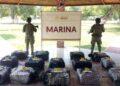 Bloque 2- Junio 4 - Especies Políticas Two Soldiers in Camouflage Stand Behind a Display of Numbered, Netted Bags on a Tiled Floor Under a Shelter, with a Marina Sign in the Background. - Especies Políticas