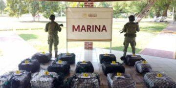 Two Soldiers in Camouflage Stand Behind a Display of Numbered, Netted Bags on a Tiled Floor Under a Shelter, with a Marina Sign in the Background. - Especies Políticas