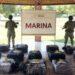 Bloque 2- Junio 4 - Especies Políticas Two Soldiers in Camouflage Stand Behind a Display of Numbered, Netted Bags on a Tiled Floor Under a Shelter, with a Marina Sign in the Background. - Especies Políticas