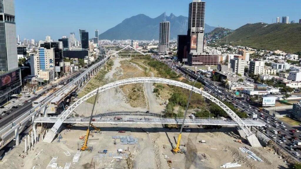 Construction Site Under a Large White Arch Bridge Spanning a Desert-like Area Between Two City Districts, with Cranes Lifting Sections and Elevated Roads Nearby. - Especies Políticas