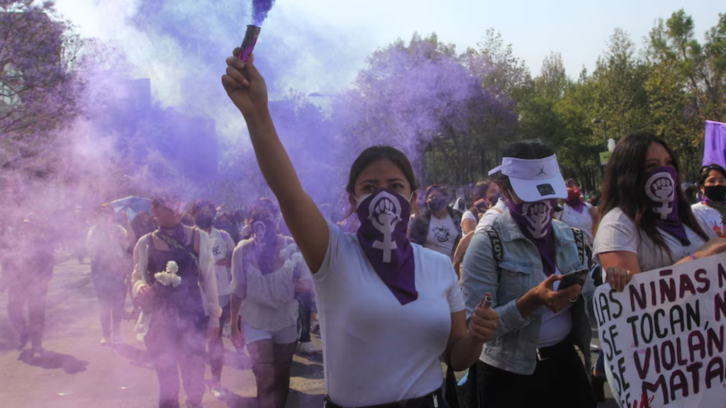 Caso-de-feminicidio - Especies Políticas Protesters March Through Smoke, a Woman in White Raises a Purple Smoke Flare with a Feminist Fist Mask Visible on Many Participants Nearby. - Especies Políticas
