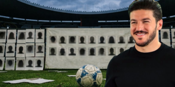 Smiling Man in a Black Shirt Standing on a Soccer Field with a Blue-and-white Soccer Ball at His Feet; Behind Him is a Wall Covered with Silhouette Mugshots. - Especies Políticas