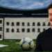 Smiling Man in a Black Shirt Standing on a Soccer Field with a Blue-and-white Soccer Ball at His Feet; Behind Him is a Wall Covered with Silhouette Mugshots. - Especies Políticas