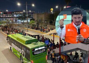 Crowd lined up at a bus terminal at night, next to a green bus; a large circular image shows a man in an orange safety vest speaking into a microphone.