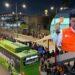 Samuel-presume-que-sobran-camiones - Especies Políticas Crowd Lined Up at a Bus Terminal at Night, Next to a Green Bus; a Large Circular Image Shows a Man in an Orange Safety Vest Speaking into a Microphone. - Especies Políticas
